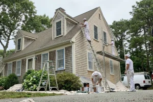 Painters apply a fresh coat of exterior paint to a shingle-style home in South Shore, Massachusetts, with ladders, drop cloths, and landscaping visible.